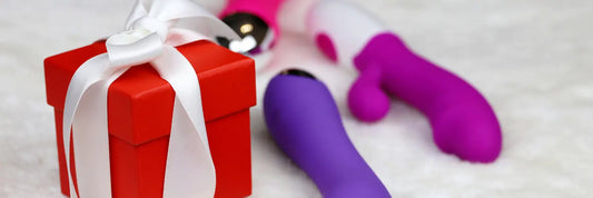 Petit coffret cadeau rouge avec un ruban blanc posé sur une surface douce, accompagné de jouets intimes colorés en arrière-plan, évoquant des idées de cadeaux coquins pour Noël. Disponible sur le love shop haut de gamme Oh My God'Z.