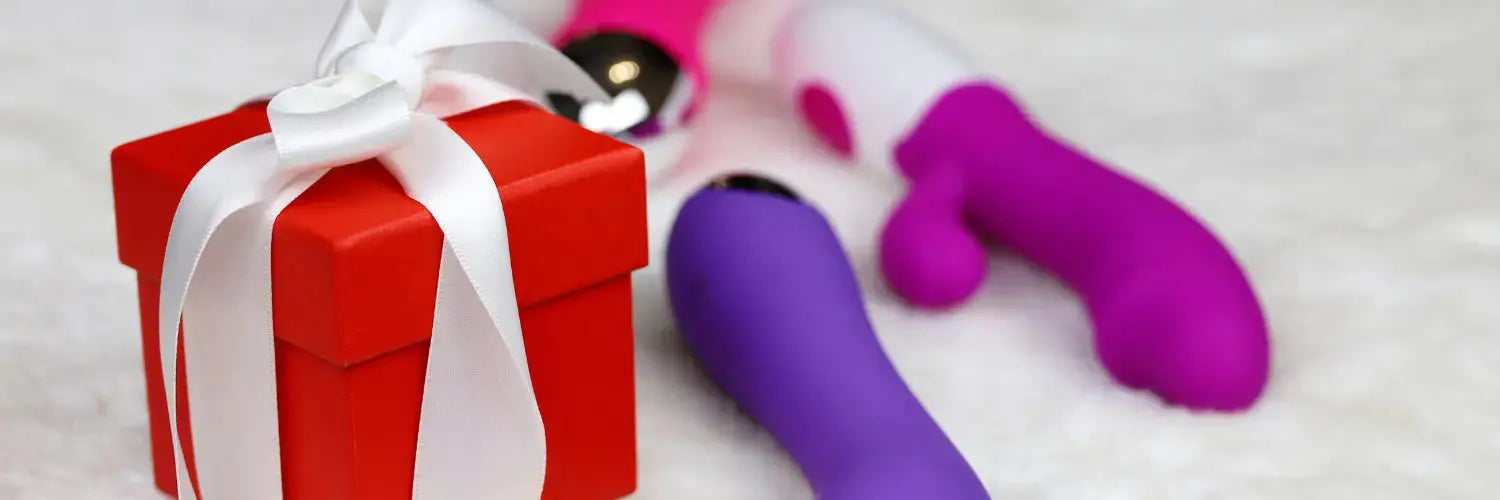 Petit coffret cadeau rouge avec un ruban blanc posé sur une surface douce, accompagné de jouets intimes colorés en arrière-plan, évoquant des idées de cadeaux coquins pour Noël. Disponible sur le love shop haut de gamme Oh My God'Z.