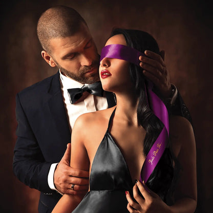 Homme et femme en situation BDSM, l'homme attache un bandeau satin pourpre Sweet Caress sur les yeux de la femme.